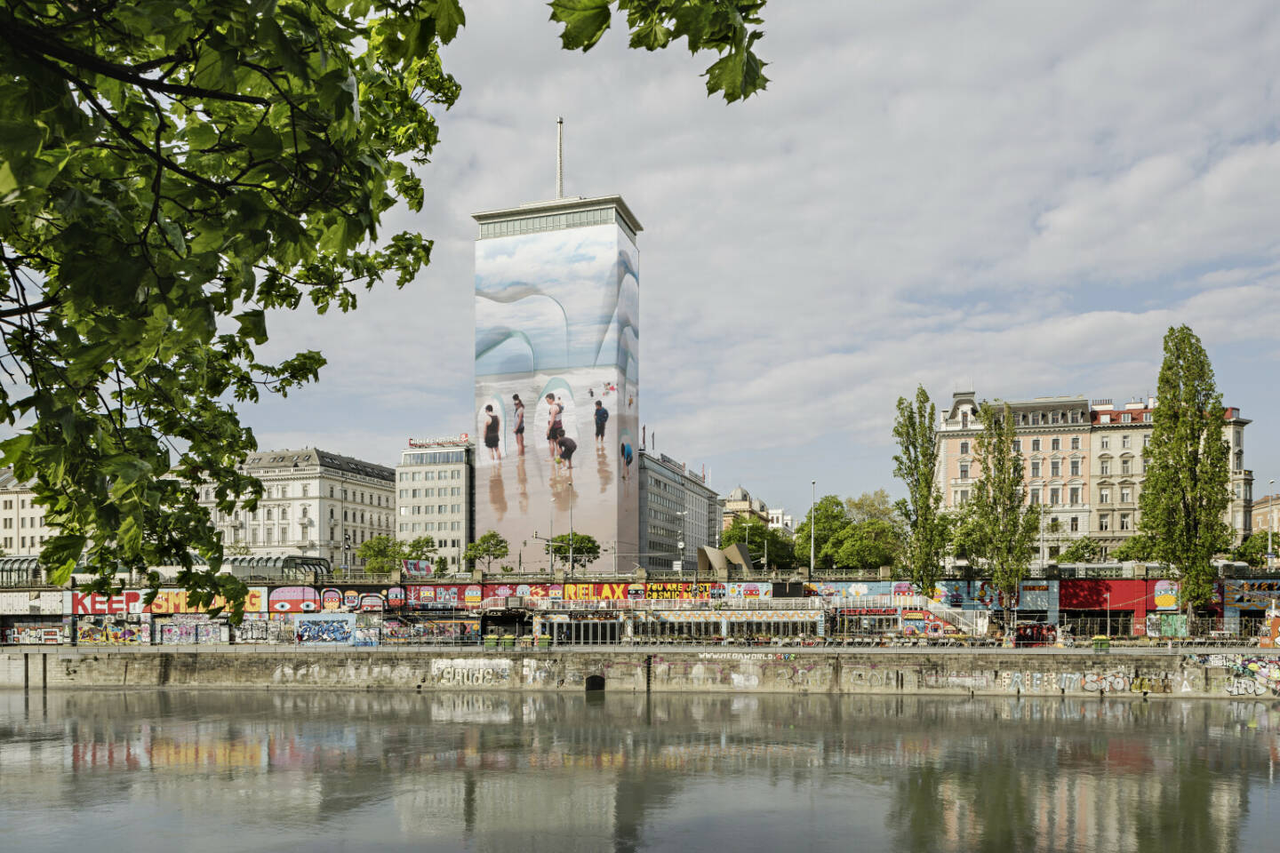 Wiener Städtische Versicherungsverein: „Wandernde Eisberge“ mitten im Wiener Stadtzentrum, Auf Initiative des Wiener Städtischen Versicherungsvereins verwandelt sich der Ringturm über die Sommermonate in eine scheinbar spielerisch sommerliche Szenerie, die Badende an einem Sandstrand zeigt. Doch die Wolken im Hintergrund und die scharfen Silhouetten, welche die slowenische Künstlerin Vanja Bućan aus dem Bild hervortreten lässt, lassen Veränderung anklingen. Credit: Wiener Städtische Versicherungsverein/Hertha Hurnaus